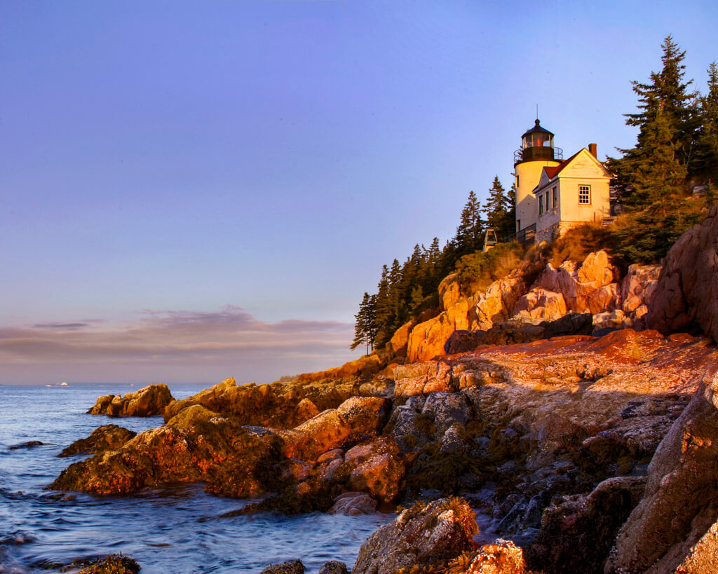 Tiny lighthouse on a cliff, above a rocky shore, with early morning sun shining on the surface at Acadia National Park.