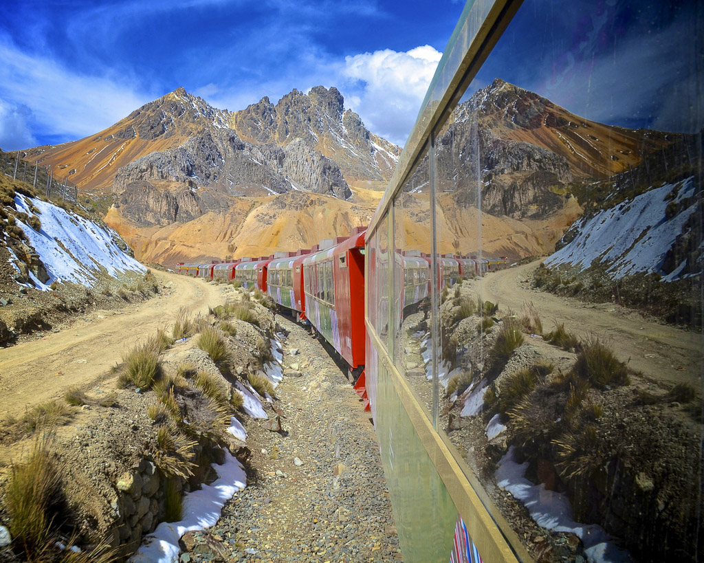 A long red train passes befre a rugged mountain, scenery from a Peru train journey.